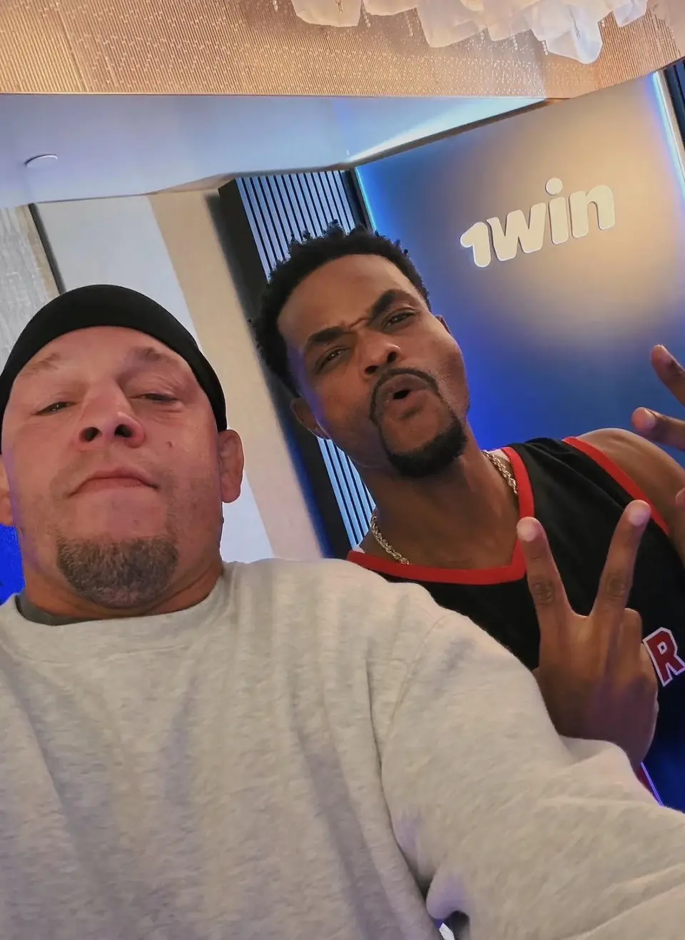 Two people taking a photo together in front of a wall with the 1win logo.